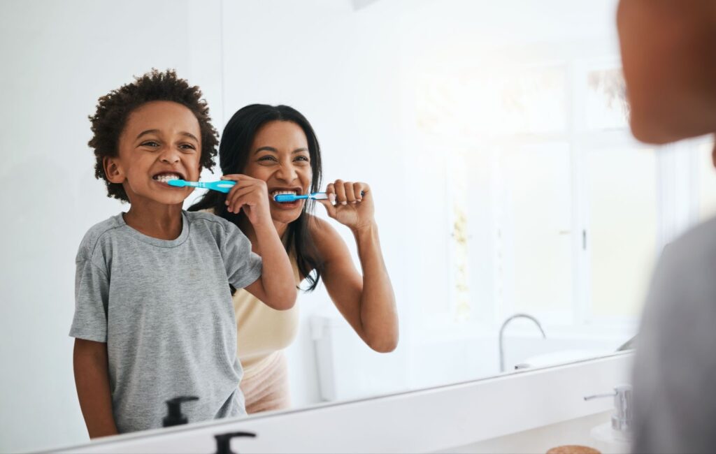 A parent and their child stand in front of a bathroom mirror together, brushing their teeth.