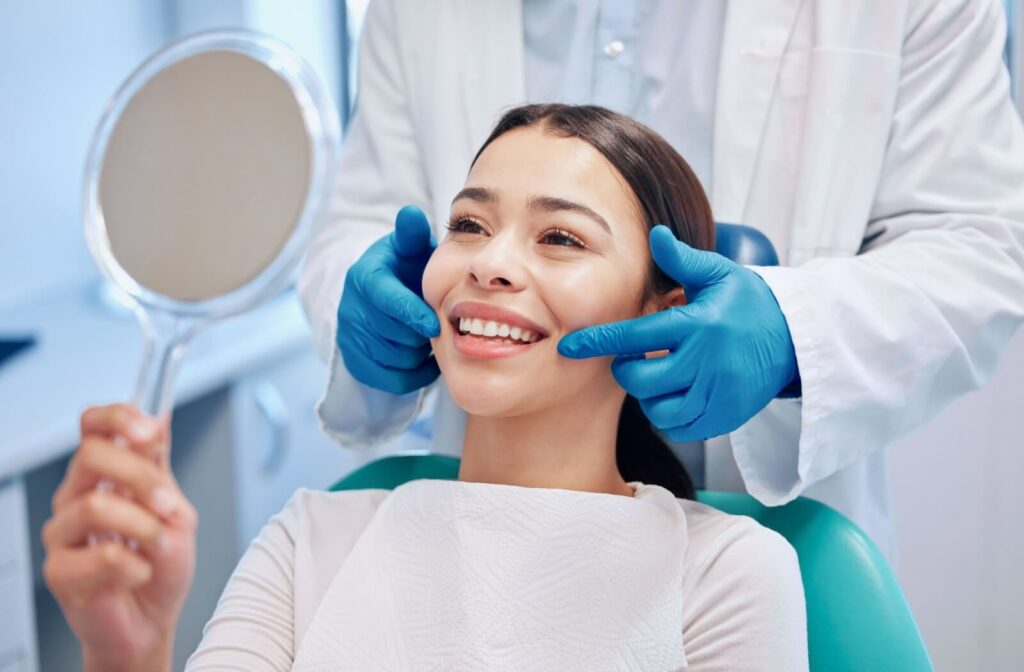 A patient smiling in a mirror while a dentist gestures to her teeth to indicate the results of replacing a dental bridge.