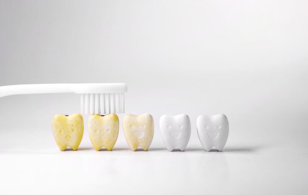 Five cartoon teeth sit on a white background. The teeth on the left are yellowed, while the teeth on the right are white. A toothbrush is cleaning the yellow teeth.