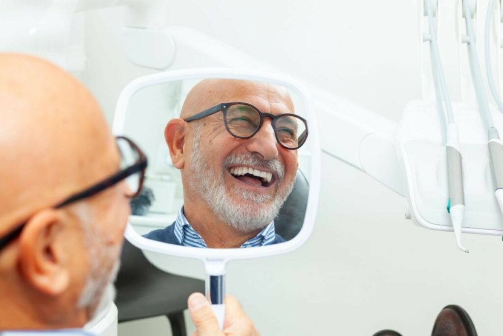 A happy senior man with a beard and glasses looking at his new smile in a handheld dental mirror while sitting in a dentist's chair at Sundance Dental.